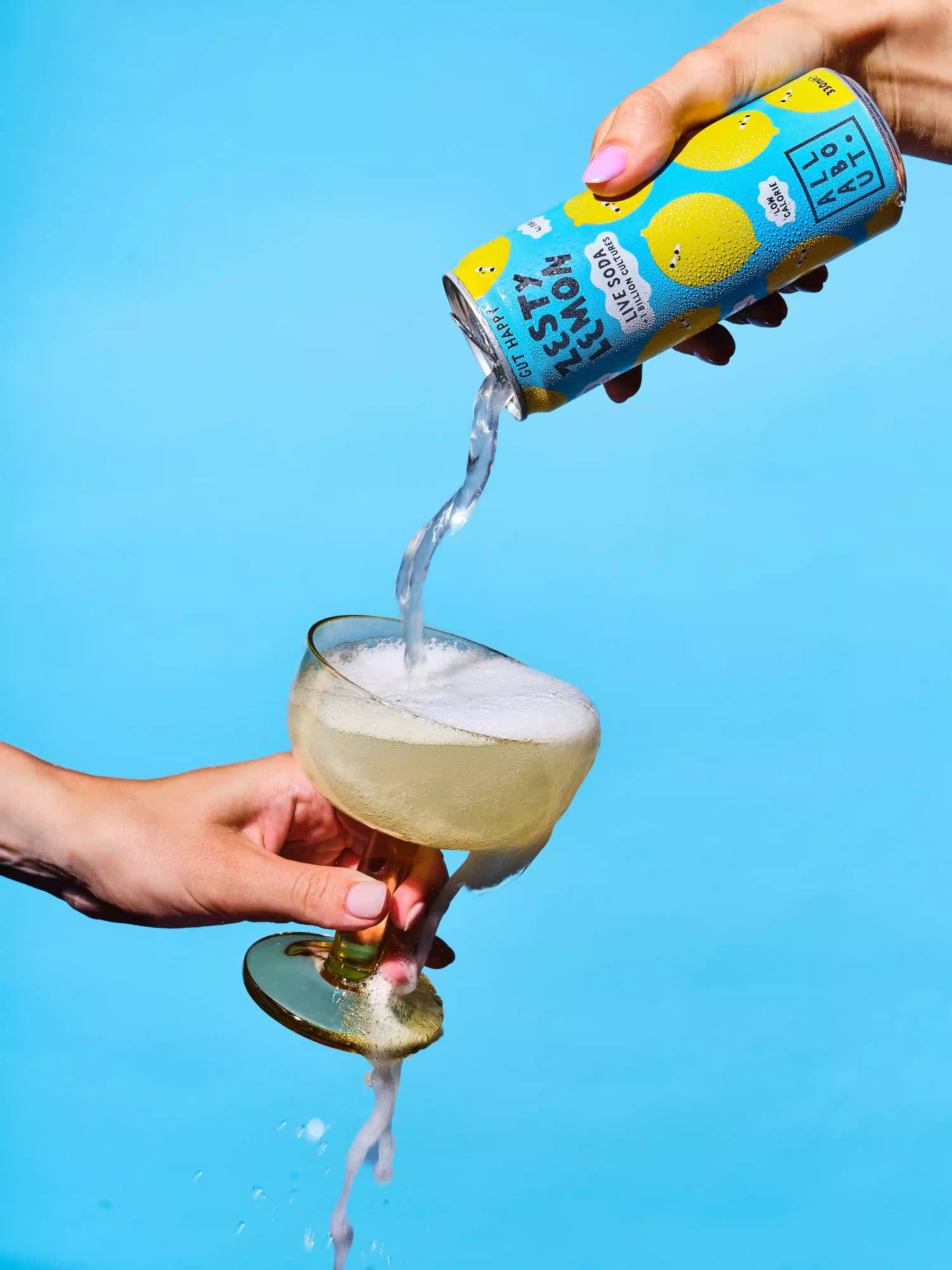 A hand holds a can of Lemon Live Soda and pours the liquid into a coupe glass filled with a pale yellow drink against a bright blue background.