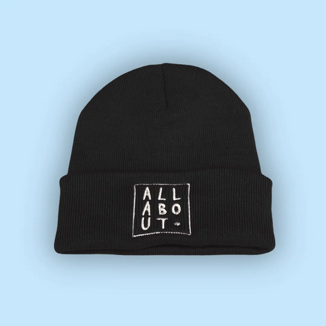 A black knit beanie hat with a folded cuff, featuring
 a white embroidered square logo on the front that 
reads 'ALL ABOUT.' in vertical letters, set against a 
solid light blue background.