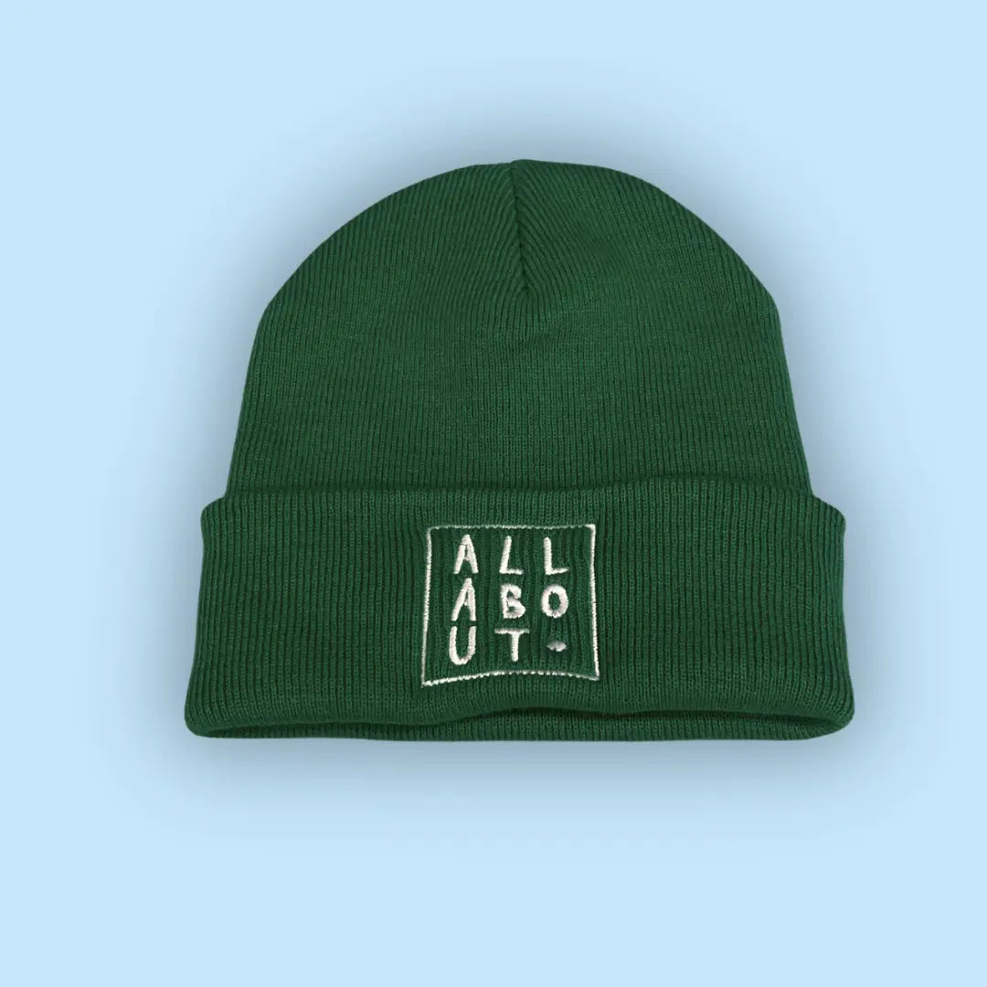 A green knit beanie hat with a folded cuff, featuring
 a white embroidered square logo on the front that 
reads 'ALL ABOUT.' in vertical letters, set against a 
solid light blue background.