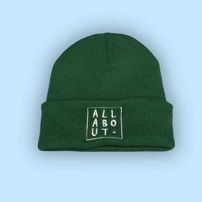 A green knit beanie hat with a folded cuff, featuring
 a white embroidered square logo on the front that 
reads 'ALL ABOUT.' in vertical letters, set against a 
solid light blue background.