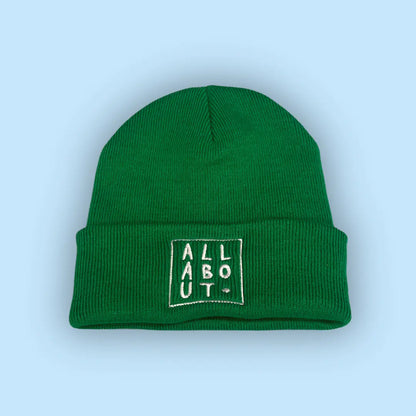 A green knit beanie hat with a folded cuff, featuring a white embroidered square logo on the front that reads 'ALL ABOUT.' in vertical letters, set against a solid light blue background.