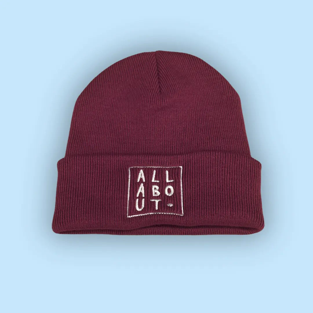 A maroon knit beanie hat with a folded cuff, featuring
 a white embroidered square logo on the front that 
reads 'ALL ABOUT.' in vertical letters, set against a 
solid light blue background.