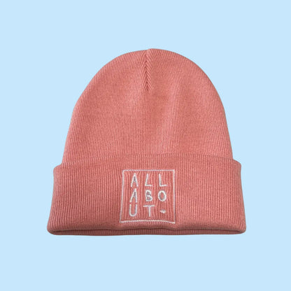 A pink knit beanie hat with a folded cuff, featuring
 a white embroidered square logo on the front that 
reads 'ALL ABOUT.' in vertical letters, set against a 
solid light blue background.
