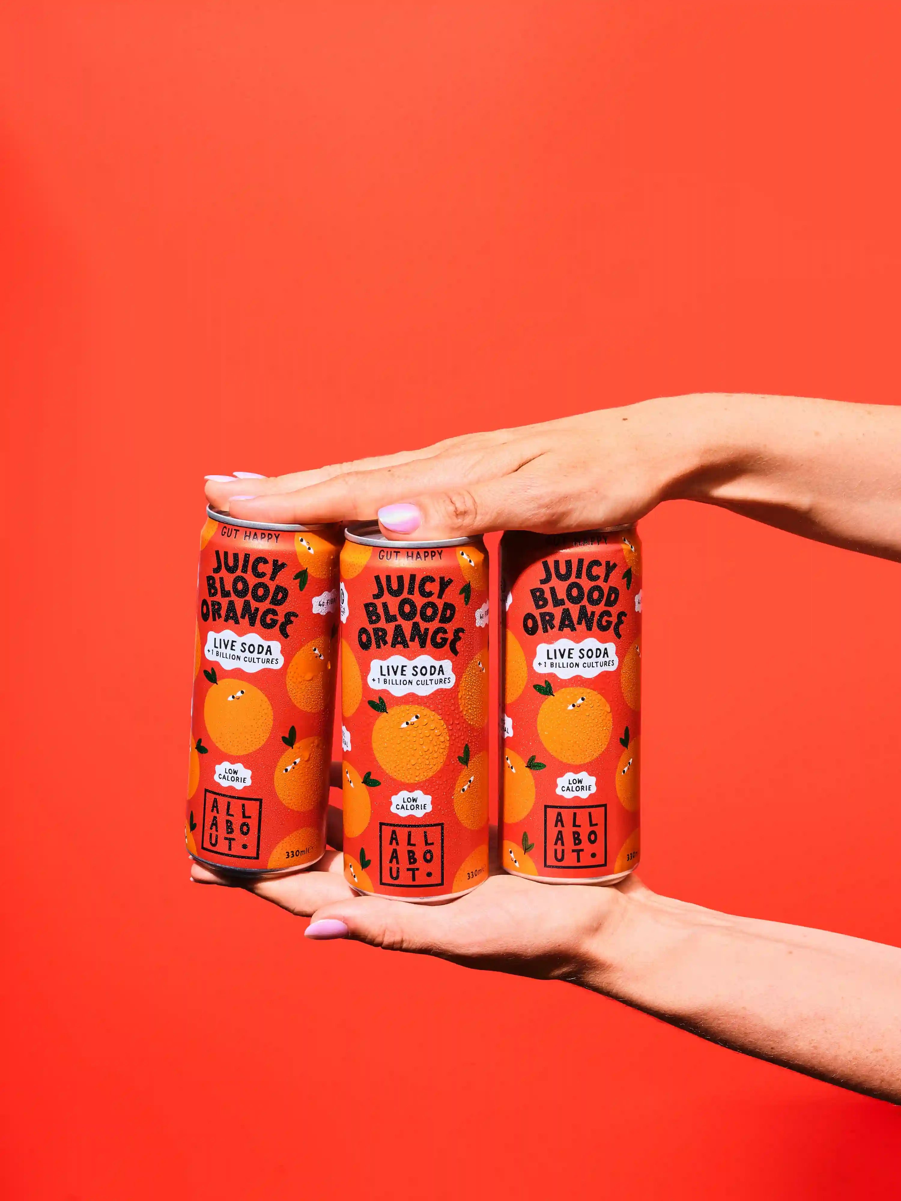 Three cans of Juicy Blood Orange Live Soda held in two hands against a solid bright red background.